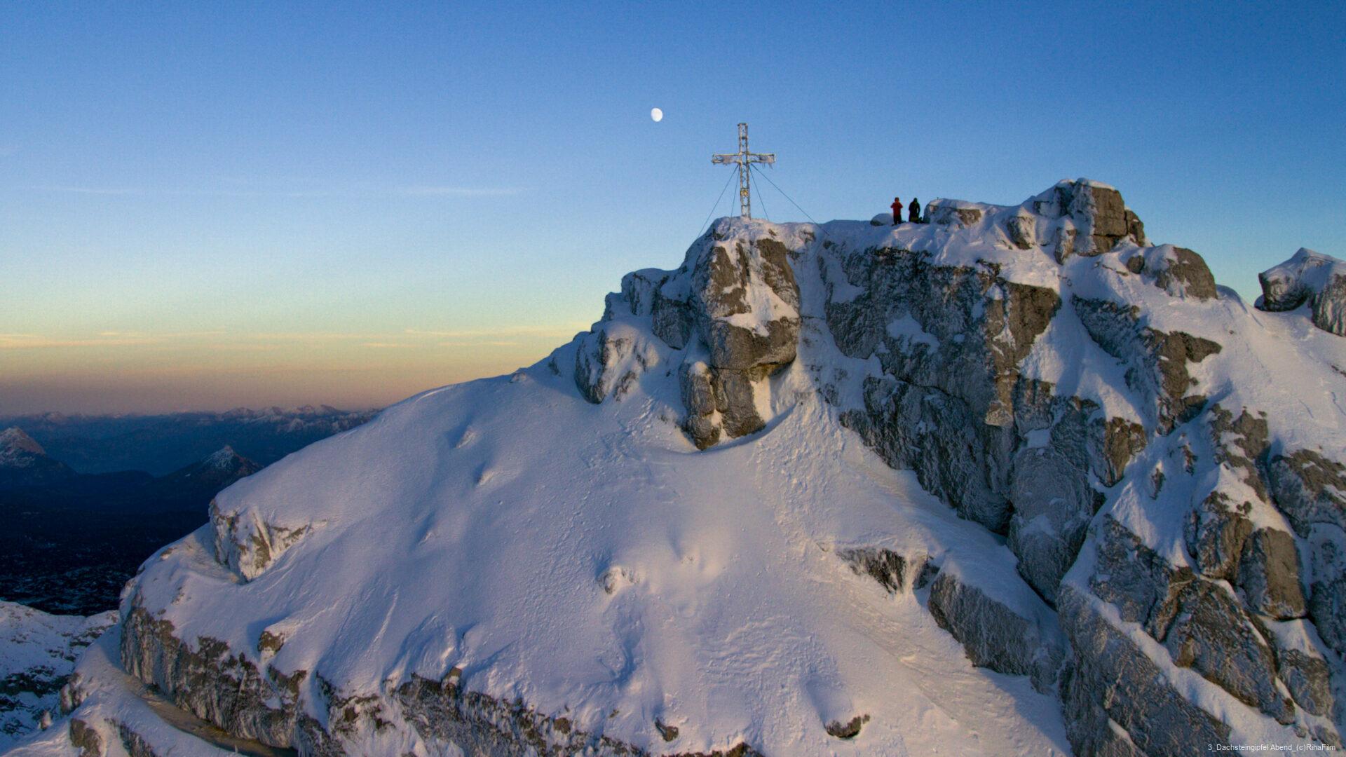 Verschneiter Berggipfel mit Gipfelkreuz, drei Personen stehen am Rand, über ihnen am Himmel der Mond.