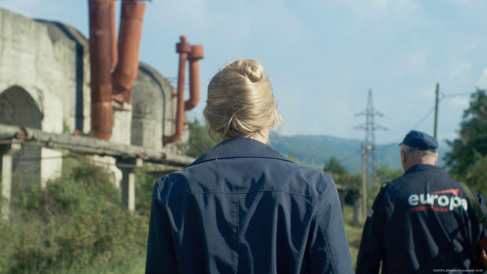 Rückansicht einer Frau mit blonden Haaren in einem Zopf und einem Mann in blauer Uniform mit der Aufschrift "europa", die in einer industriellen Umgebung mit Betongebäuden und roten Rohren spazieren gehen.
