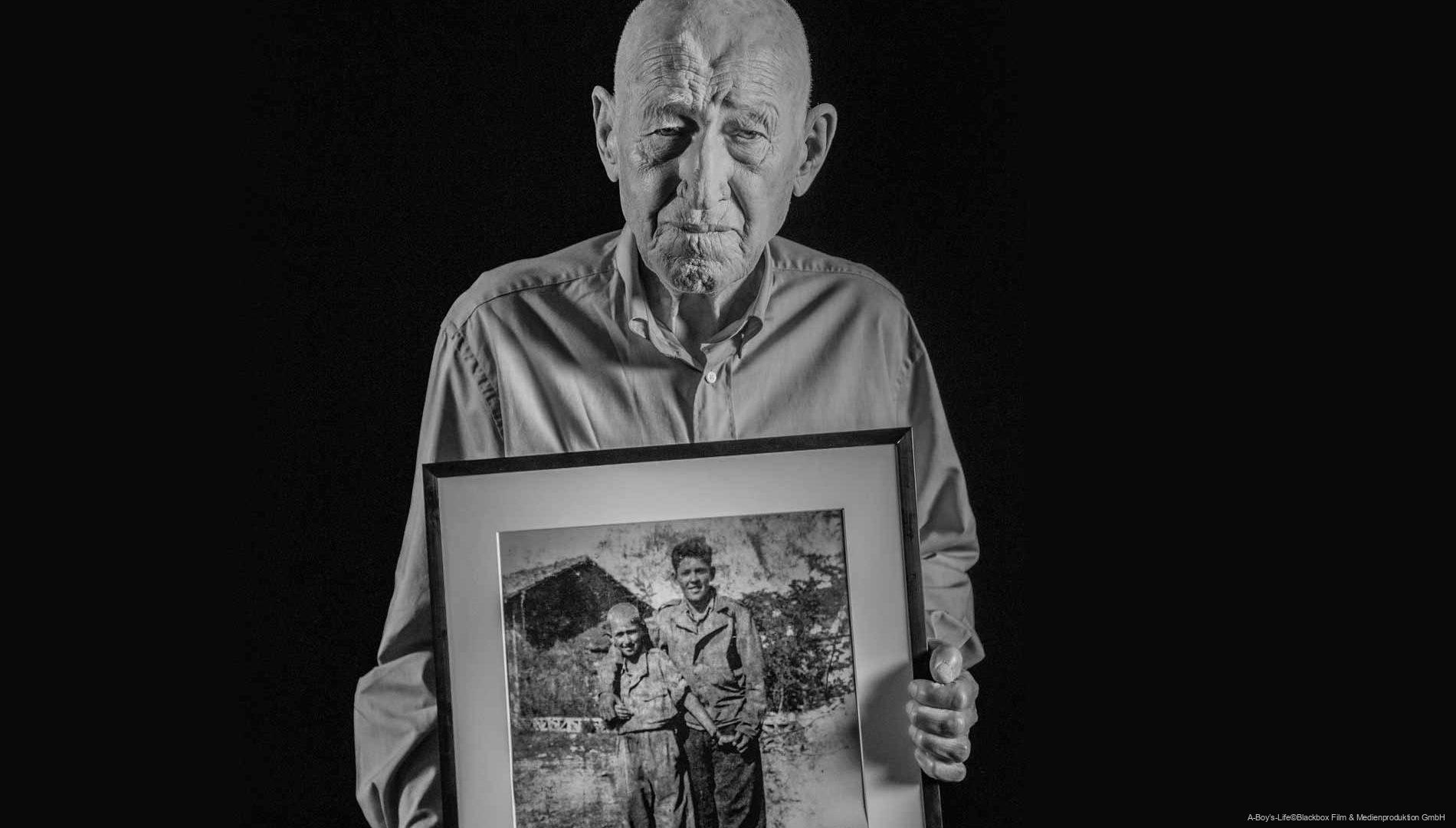 Älterer Mann in Hemd vor schwarzem Hintergrund, der einen gerahmten, alten Schwarzweiß-Fotodruck mit zwei Jungen hält.