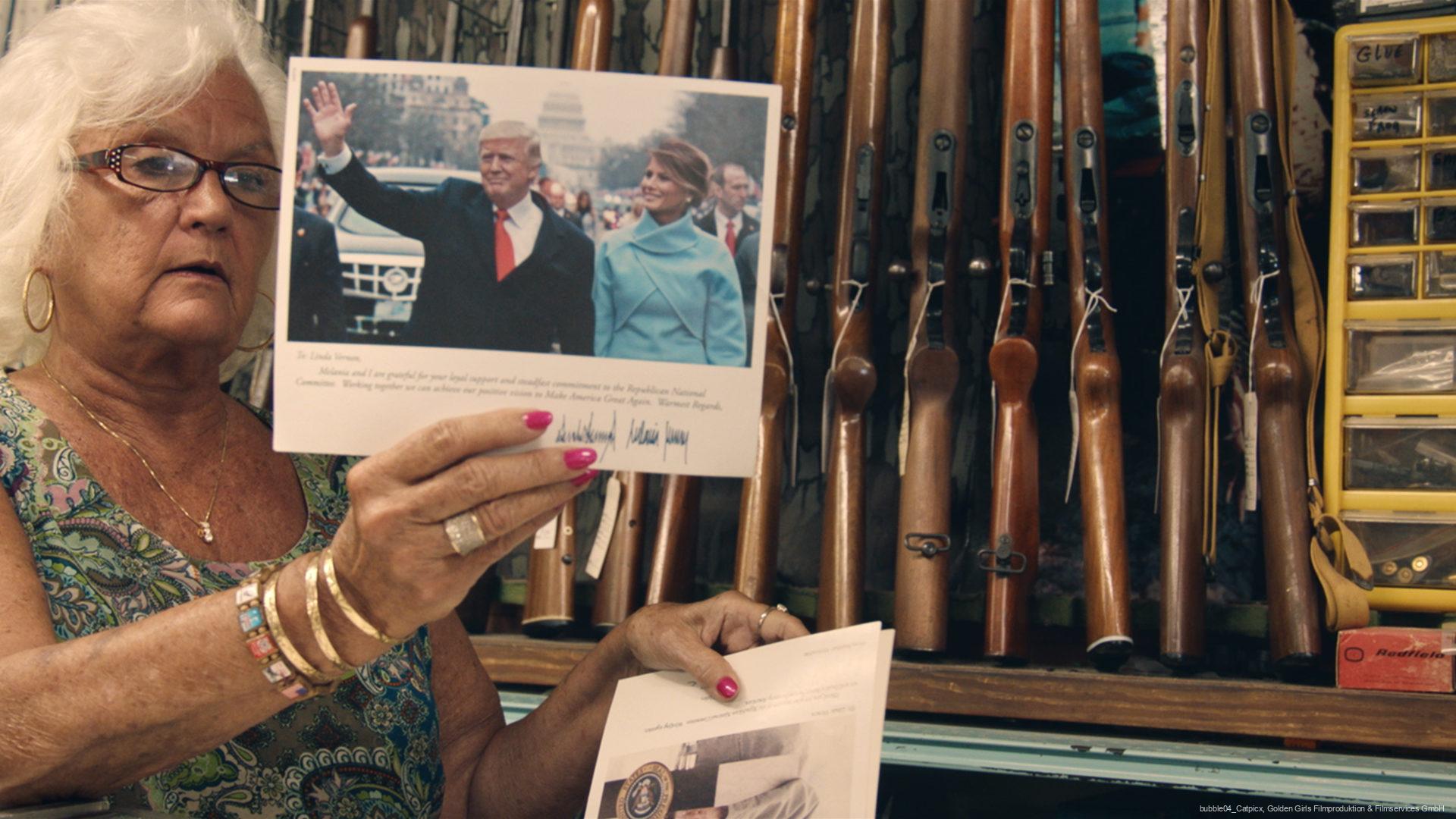 Ältere Frau mit weißem Haar, Brille und buntem Oberteil hält ein Foto von Donald Trump und Melania Trump in einem Raum mit an der Wand stehenden Gewehren und einem gelben Organizer für Kleinteile.