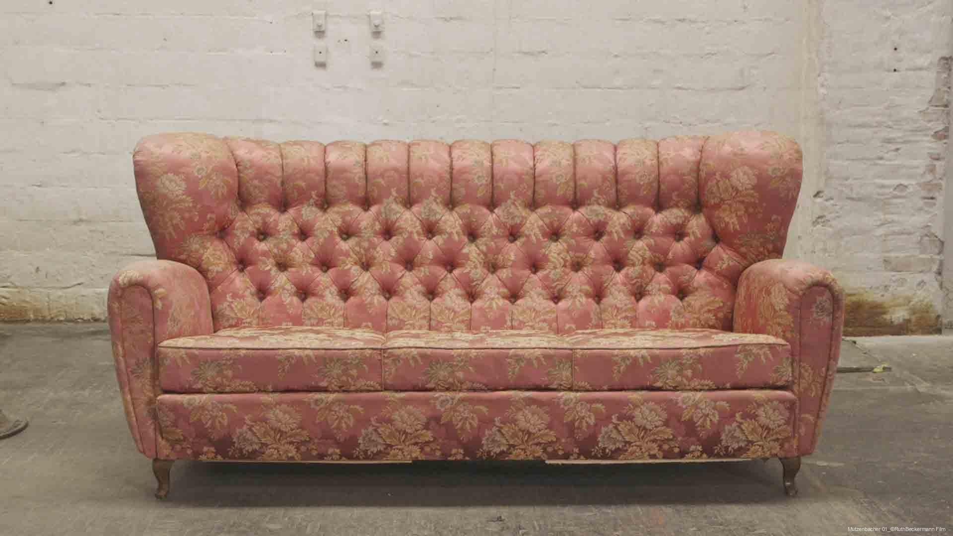 Rosa Vintage-Sofa mit floralen Mustern und gepolstertem Rückenlehnen- und Armlehnenbereich vor einer weißen Backsteinwand auf grauem Betonboden