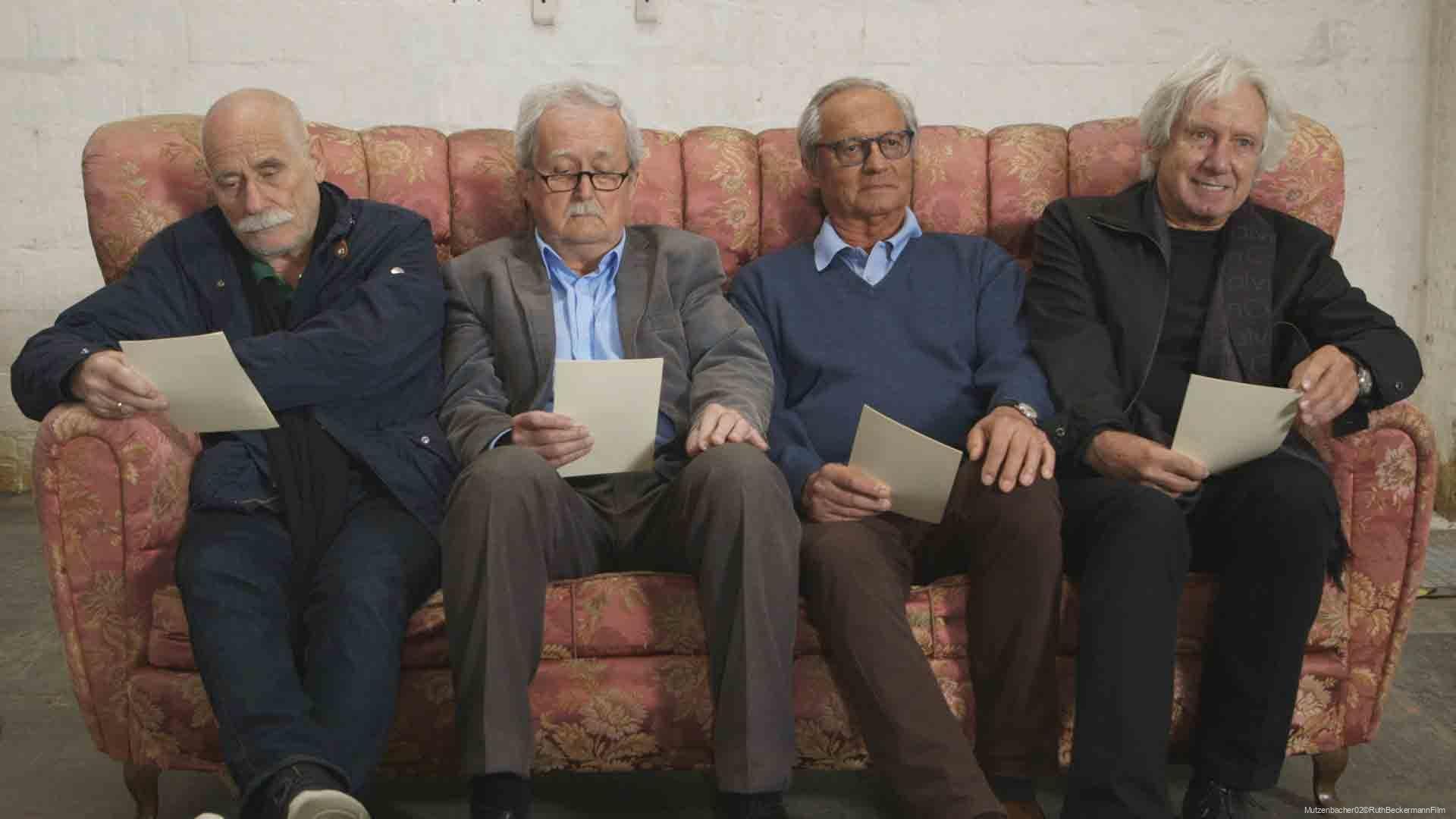 Vier ältere Männer sitzen nebeneinander auf einem altrosa gemusterten Sofa, jeder hält ein Blatt Papier in der Hand, der erste und zweite von links tragen Brillen, der vierte Mann lächelt.