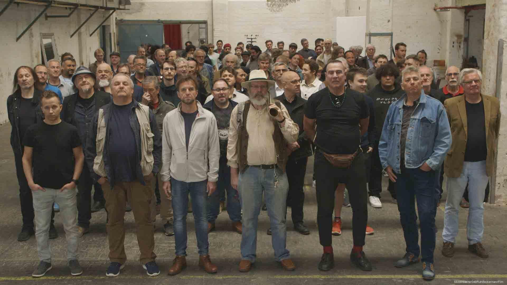 Große Gruppe von Männern unterschiedlichen Alters, die in einer alten industriellen Halle stehen und in die Kamera schauen, der vordere Mann trägt einen Hut und hält ein Fernglas über der Schulter.