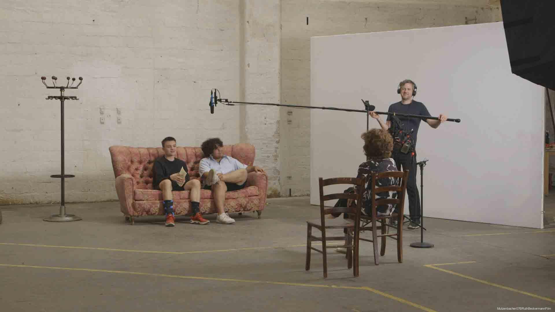 Zwei Männer sitzen auf einem rosa Sofa in einem großen, hellen Raum, während eine Frau auf einem Holztstuhl vor einer weißen Wand sitzt und ein Mann mit Kopfhörern am Tonaufnahme-Mikrofon arbeitet, das auf einer Auslegerstange gehalten wird.