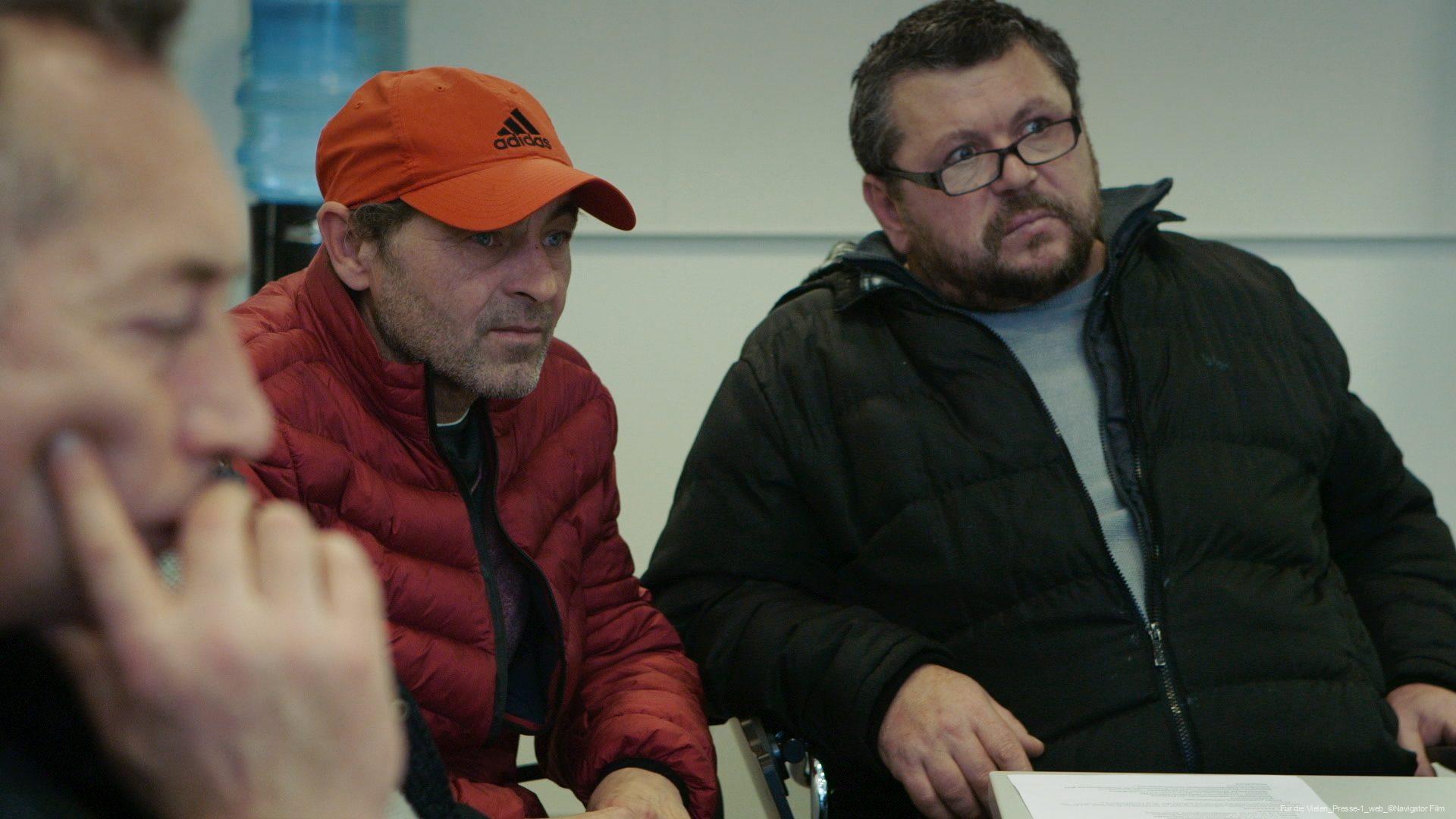 Drei Männer in Winterjacken sitzen zusammen an einem Tisch in einem Innenraum, einer trägt eine rote Adidas-Kappe, ein anderer eine schwarze Jacke und Brille, der dritte ist verschwommen im Vordergrund.