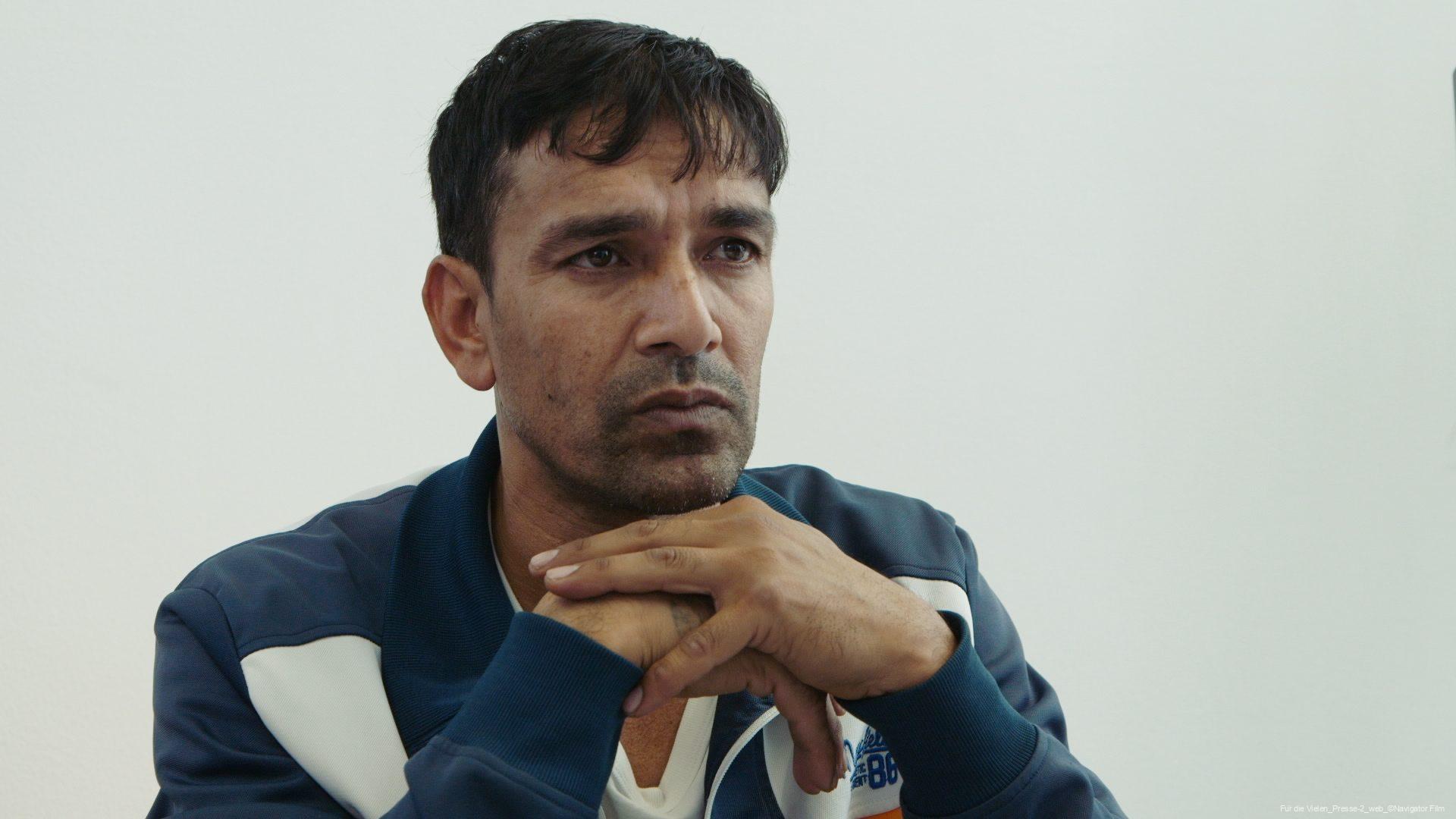 Mann mit kurzem dunklem Haar und dunkelblauen Jacke, der nachdenklich mit verschränkten Händen vor dem Gesicht in die Ferne schaut, vor einem hellen Hintergrund