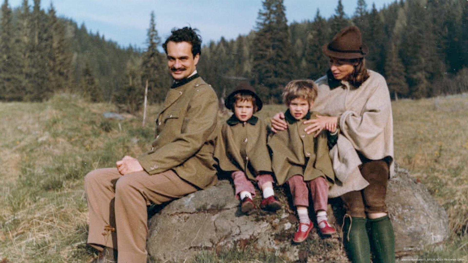 Eine Familie mit zwei Erwachsenen und zwei Kindern sitzt auf einem Felsen in einer Wiesenlandschaft vor einem Wald, alle tragen traditionelle Kleidung in Erdtönen, die Mutter hat einen Hut und grüne Stiefel an.