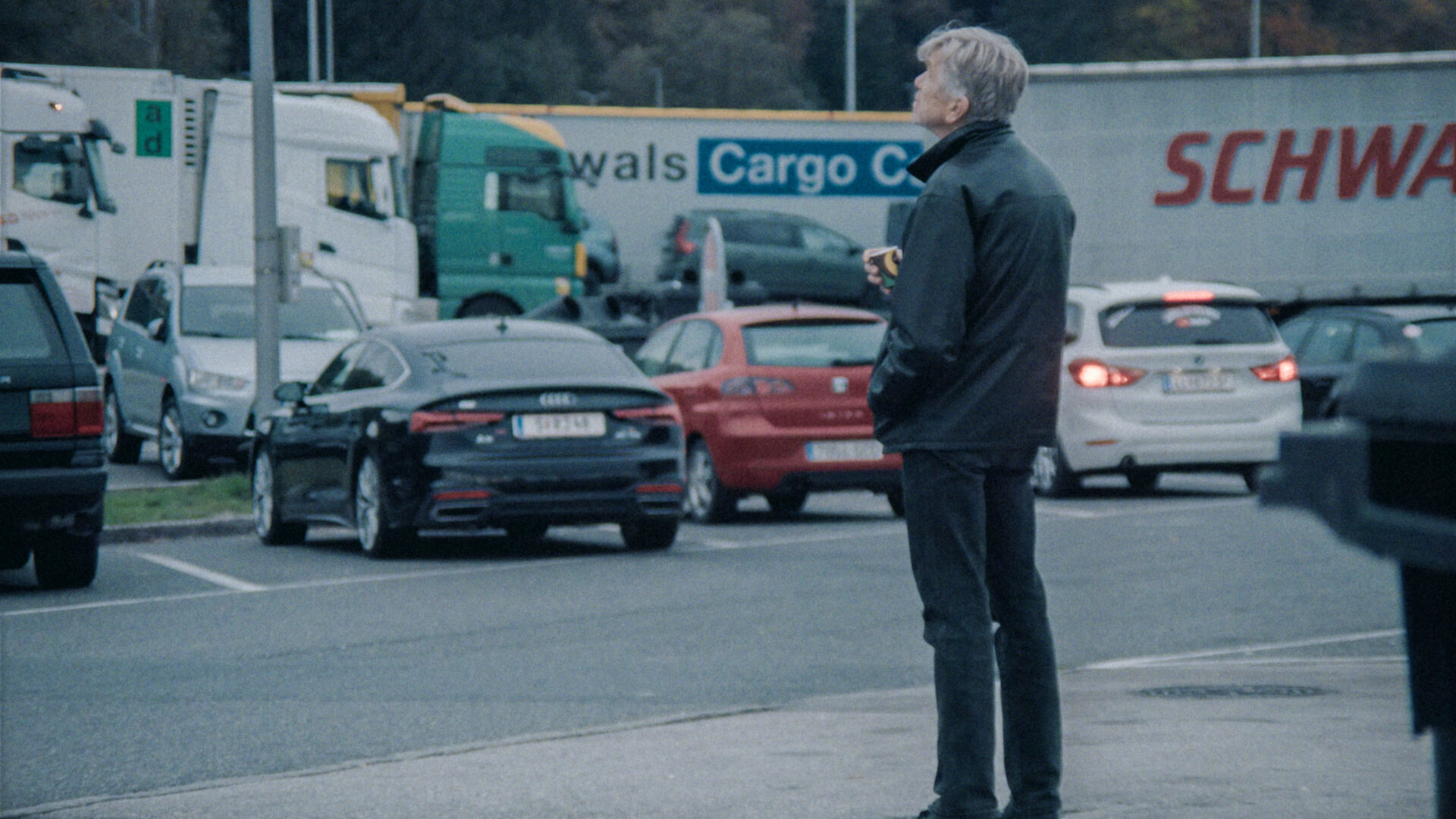 Älterer Mann steht zwischen wartenden Autos und Lastwagen auf einem Parkplatz