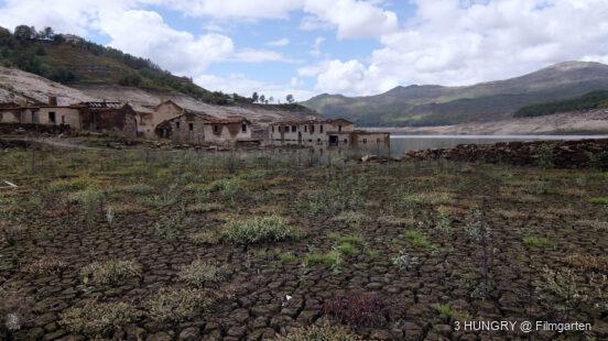 Verlassene Häuser an einem ausgetrockneten See, Risse im Boden und vereinzelte Pflanzen, umgeben von Bergen und Wolken.