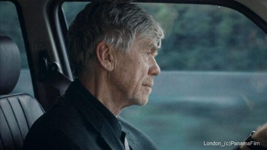 Älterer Mann mit grauen Haaren sitzt konzentriert am Steuer eines Autos, im Hintergrund ist eine verschwommene grüne Landschaft zu sehen.