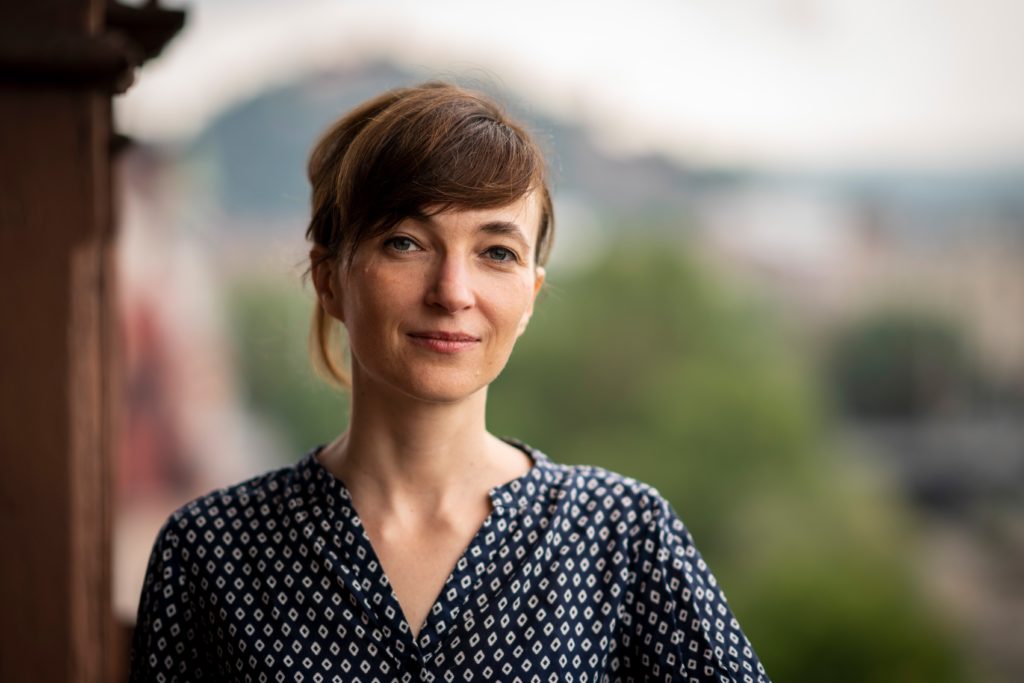 Porträt einer lächelnden Frau mit braunen Haaren, die einen dunkelblauen Pullover mit weißem Muster trägt, vor unscharfem grünen und städtischen Hintergrund.