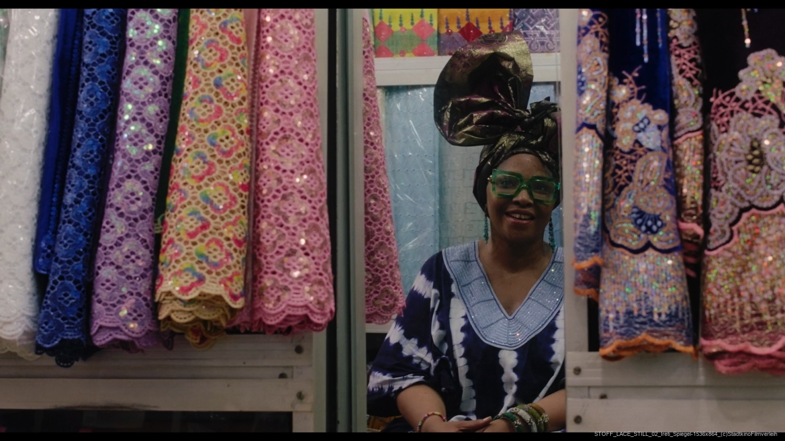 Eine Frau in traditioneller bunter Kleidung und großer Kopfbedeckung sitzt zwischen Stoffrollen mit aufwendigen Mustern.