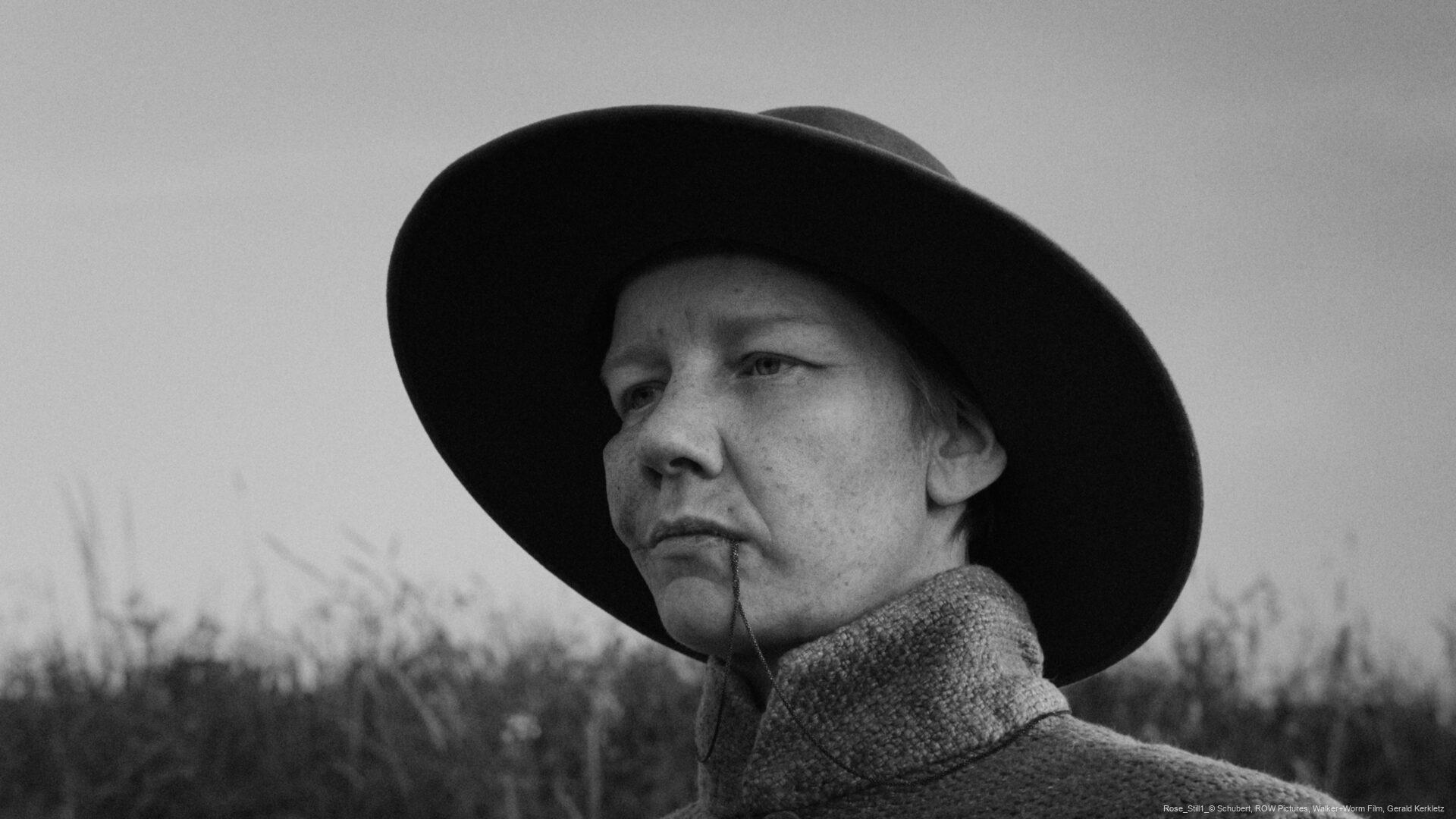 Person mit breitem Hut und ernstem Gesichtsausdruck steht im Freien vor einem unscharfen Hintergrund mit hohem Gras.