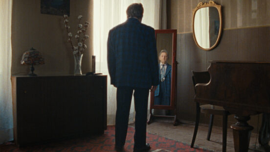 Ein Mann steht mit dem Rücken zur Kamera vor einem Spiegel, in dem sein Gesicht und seine Vorderseite sichtbar sind, in einem spärlich eingerichteten Zimmer.