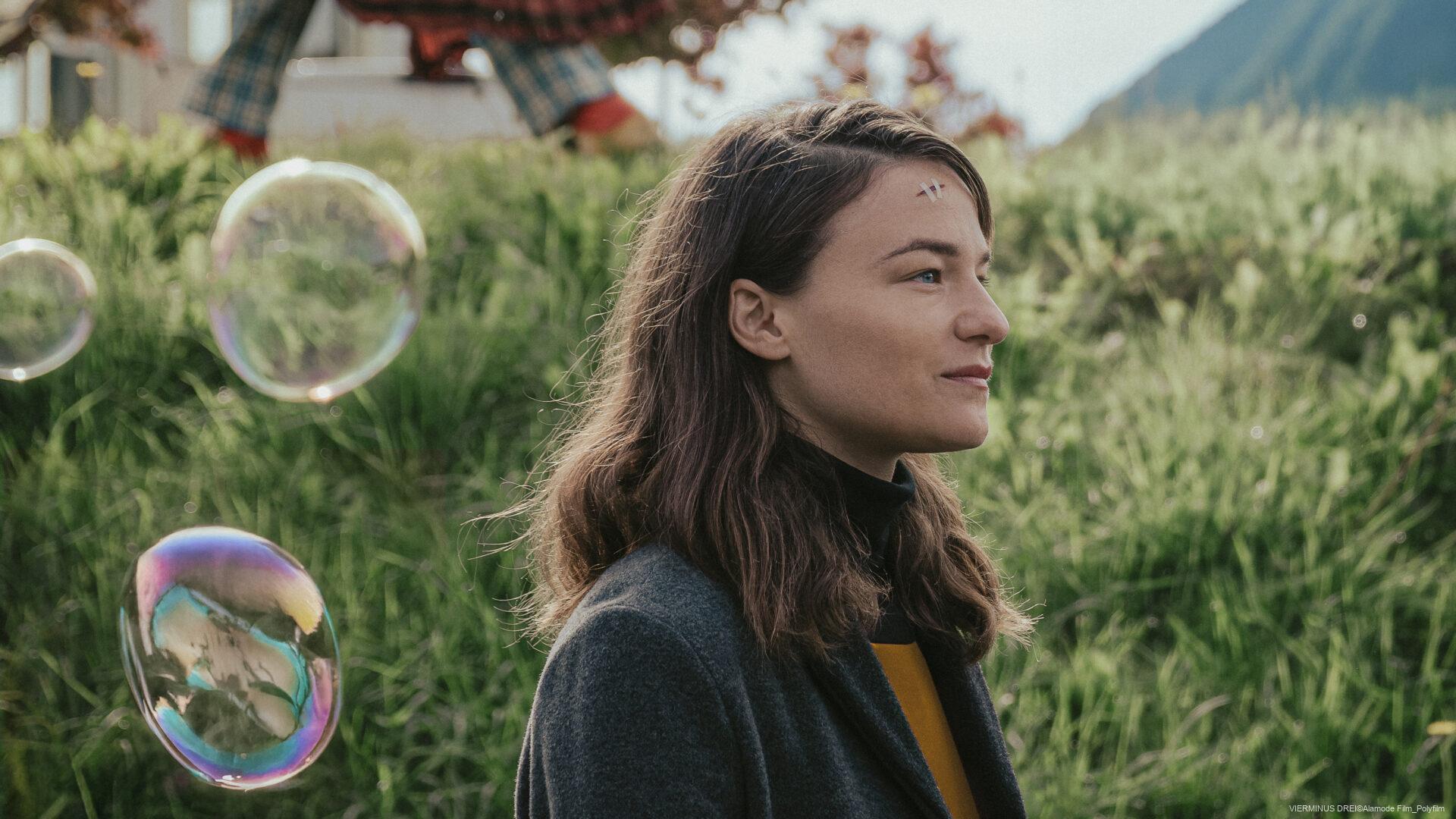 Junge Frau mit braunen Haaren steht im Profil auf einer Wiese, Seifenblasen schweben in der Luft, im Hintergrund erkennt man grüne Büsche und Berge.