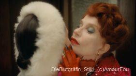 Eine elegant gekleidete Frau mit rotem Haar und leuchtendem Make-up hält das Kinn einer anderen Person mit weißem Pelzkragen fest.