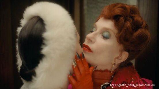 Eine elegant gekleidete Frau mit rotem Haar und leuchtendem Make-up hält das Kinn einer anderen Person mit weißem Pelzkragen fest.