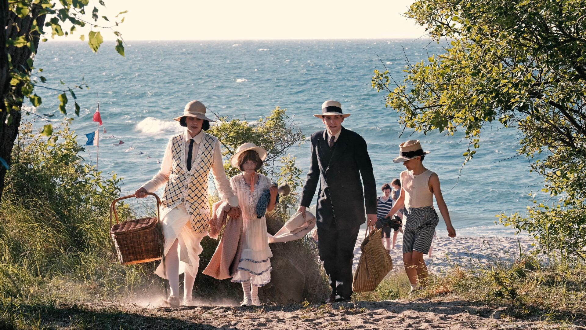 Vier Personen in historischer Kleidung mit Hüten gehen ausgehend vom Strand durch einen sandigen, bewachsenen Pfad entlang der Küste, im Hintergrund Meer und weitere Spaziergänger.