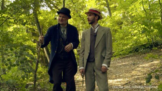 Zwei Männer in historischer Kleidung gehen in einem grünen Wald, einer trägt einen Gehstock und einen schwarzen Hut, der andere einen Anzug mit Weste und einen Strohhut mit rotem Band, letzterer hält eine Pistole in der rechten Hand.