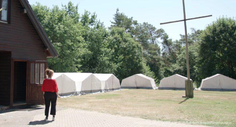 Person in rotem Oberteil und schwarzer Hose steht vor Holzhütte mit offener Tür, im Hintergrund mehrere weiße Zelte auf Rasenfläche neben Bäumen und einem einfachen Holzkreuz.