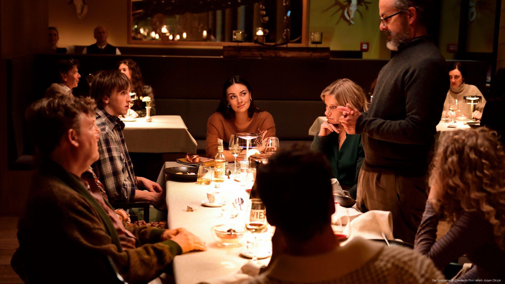 Menschen sitzen um einen festlich gedeckten Tisch in einem Restaurant, während ein Mann stehend spricht und die anderen aufmerksam zuhören; die Atmosphäre ist warm und einladend mit indirektem Licht und Dekorationen an den Wänden.