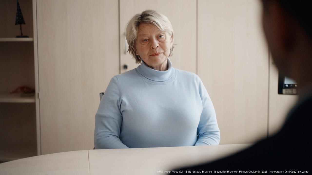 Ältere Frau mit blond-grauem Haar und hellblauem Pullover sitzt an einem Tisch und blickt eine Person gegenüber an.