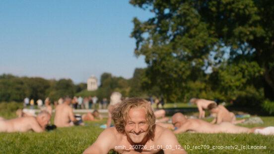 Ein lächelnder Mann liegt nackt auf einer Wiese, im Hintergrund entspannen weitere Menschen im Park bei Sonnenschein.