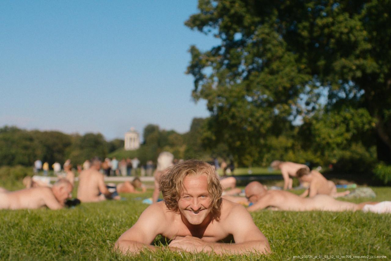 Ein lächelnder Mann liegt nackt auf einer Wiese, im Hintergrund entspannen weitere Menschen im Park bei Sonnenschein.