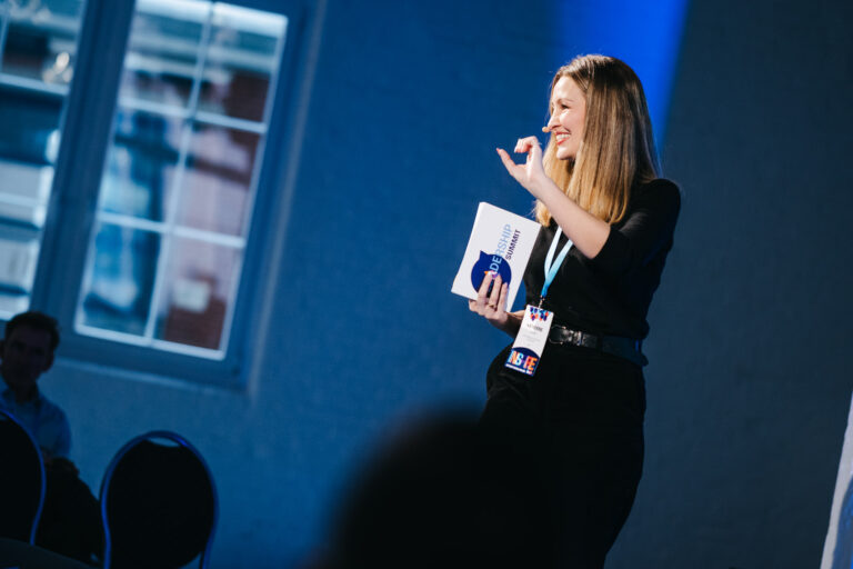 Frau mit langen blonden Haaren hält ein Notizbuch mit der Aufschrift "Leadership Summit" und spricht auf einer Bühne, während sie ein Handzeichen macht, im Hintergrund unscharfe Personen und ein Fenster.
