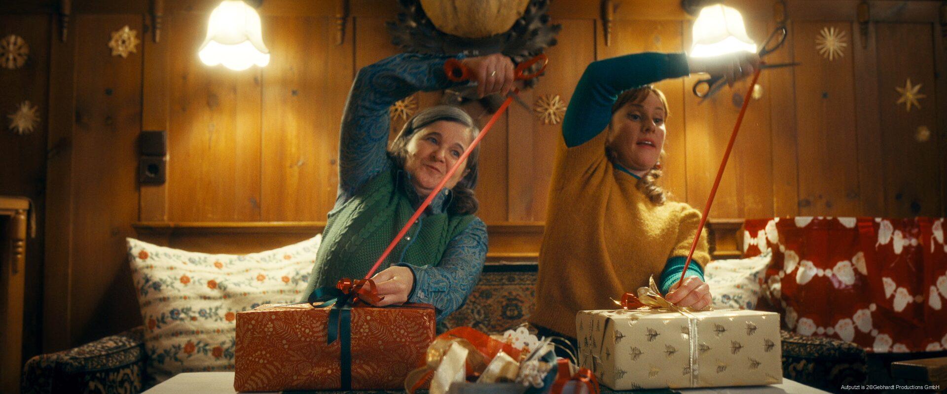 Zwei Frauen sitzen in einer gemütlichen, holzgetäfelten Stube und verpacken Geschenke, während sie gleichzeitig Geschenkband mit einer Schere schneiden; im Hintergrund sind weihnachtliche Dekorationen und warme Beleuchtung zu sehen.