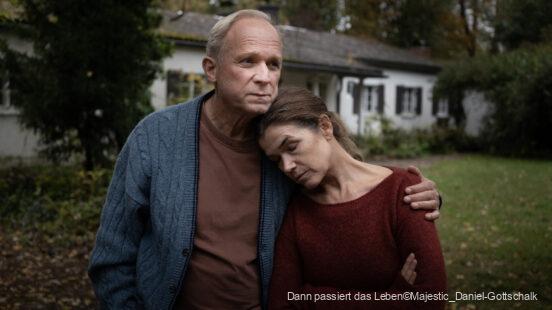 Ein älteres Paar steht eng umschlungen im Garten vor einem Haus, die Frau lehnt ihren Kopf traurig an die Schulter des Mannes, beide wirken nachdenklich und besorgt.