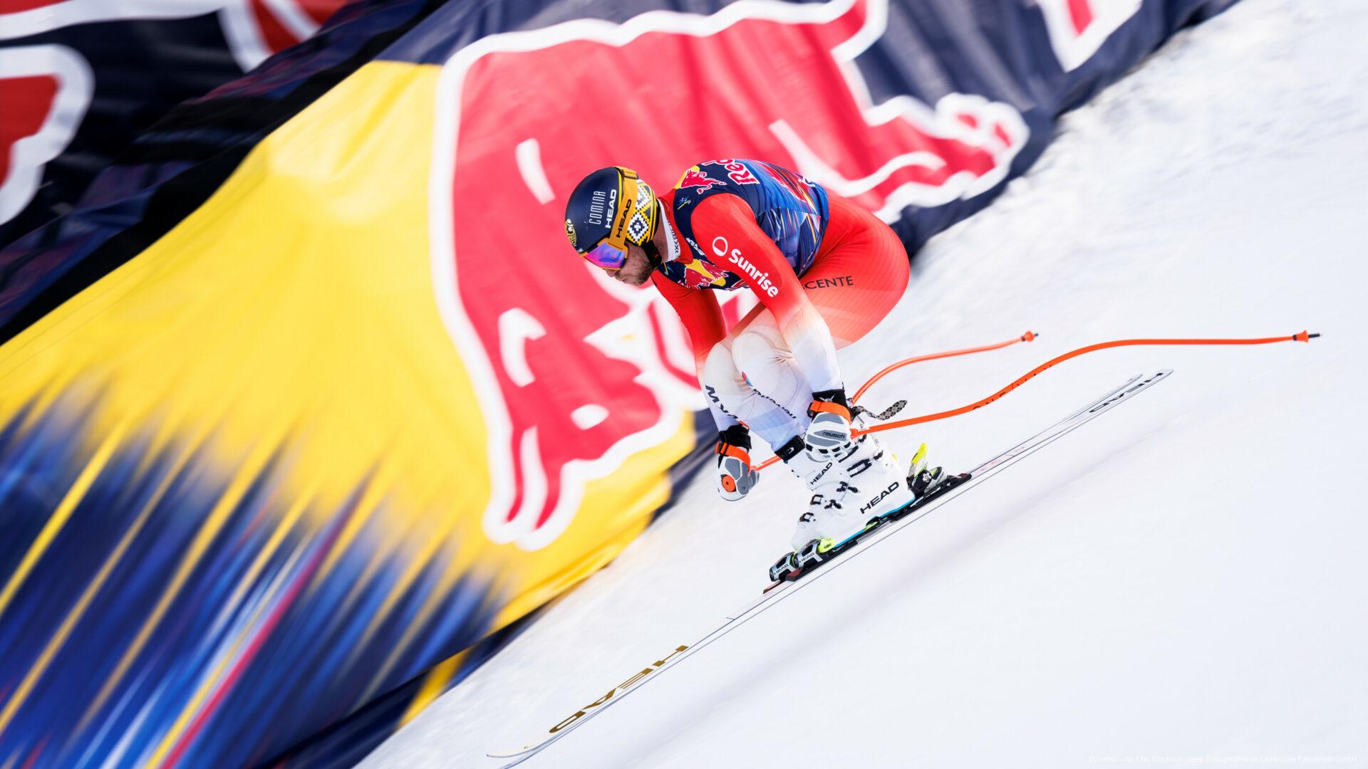 Skirennläufer in voller Abfahrtshaltung auf einer schneebedeckten Piste mit auffälligem Red Bull Banner im Hintergrund.