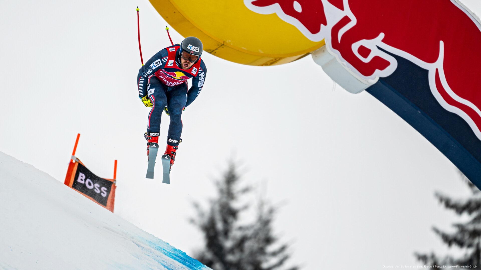 Skirennfahrer in der Luft während eines alpinen Rennlaufs, unter einem großen Red-Bull-Tor, mit Schnee, blassem Himmel und Bäumen im Hintergrund.
