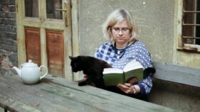 Frau mit blonden Haaren und Brille sitzt auf einer Holzbank neben einer Hauswand, liest ein Buch und streichelt dabei eine schwarze Katze; auf dem Tisch steht eine Teekanne.