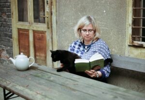 Frau mit blonden Haaren und Brille sitzt auf einer Holzbank neben einer Hauswand, liest ein Buch und streichelt dabei eine schwarze Katze; auf dem Tisch steht eine Teekanne.
