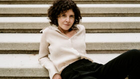 Frau mit lockigen dunklen Haaren sitzt entspannt auf einer Steintreppe, trägt eine helle Bluse und dunkle Hose.