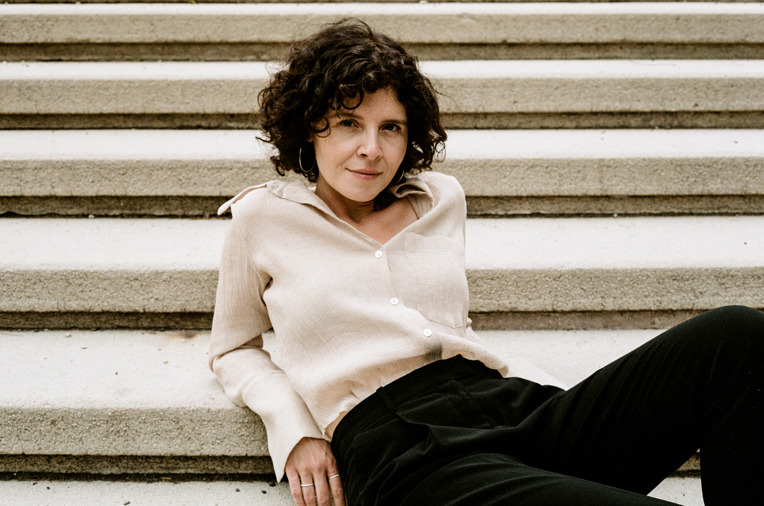 Frau mit lockigen dunklen Haaren sitzt entspannt auf einer Steintreppe, trägt eine helle Bluse und dunkle Hose.