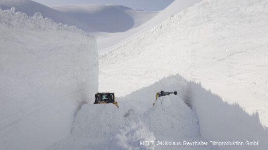 Zwei Schneepflüge arbeiten zwischen hohen, gestapelten Schneewänden in einer tief eingeschneiten Landschaft.