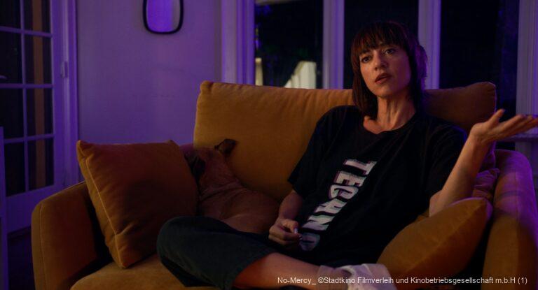 Frau mit dunklem Haar sitzt abends auf einem gelben Sofa, trägt ein schwarzes T-Shirt mit dem Schriftzug "TECHNO" und hebt fragend die Hand; neben ihr liegt ein brauner Hund, der sich ins Kissen kuschelt.