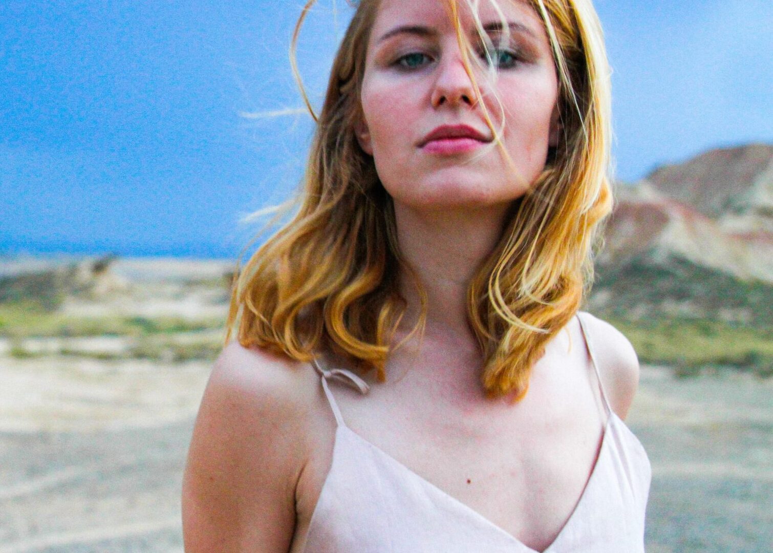 Junge Frau mit blondem Haar und hellrosa Top steht in einer trockenen, offenen Landschaft unter einem bewölkten, blauen Himmel.