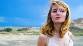 Junge Frau mit blondem, windverwehtem Haar steht in heller Sommerkleidung vor einer trockenen, weiten Landschaft unter einem dramatisch blauen Himmel.