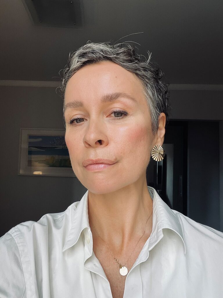 Frau mit kurzen, leicht grau melierten Haaren, weißer Bluse, goldfarbener Halskette und auffälligem Ohrring, vor neutralem Hintergrund.