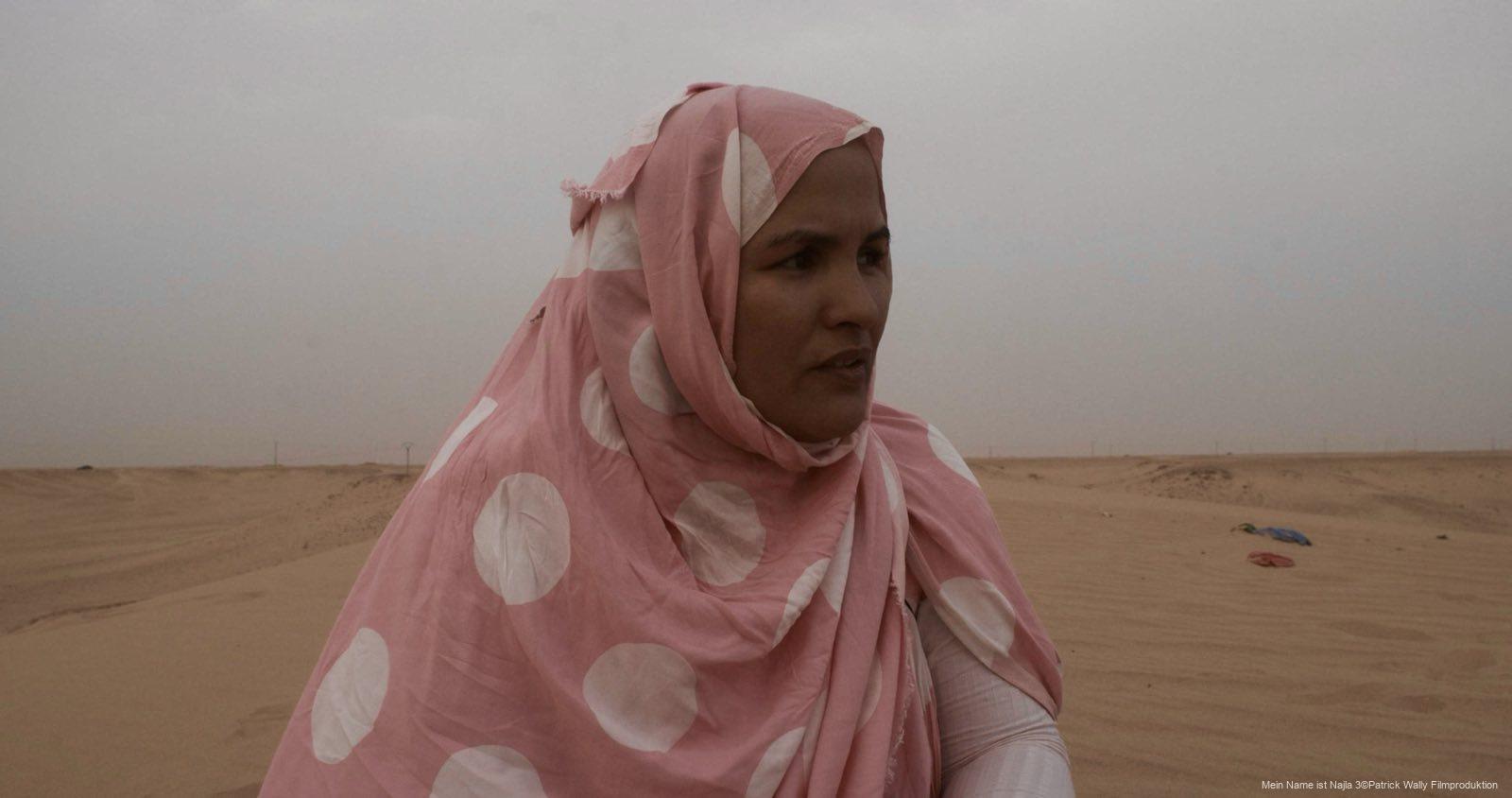 Frau in rosa Kleidung mit weißen Punkten, deren Kopf mit einem Tuch bedeckt ist, steht in einer sandigen Wüstenlandschaft unter wolkigem Himmel.