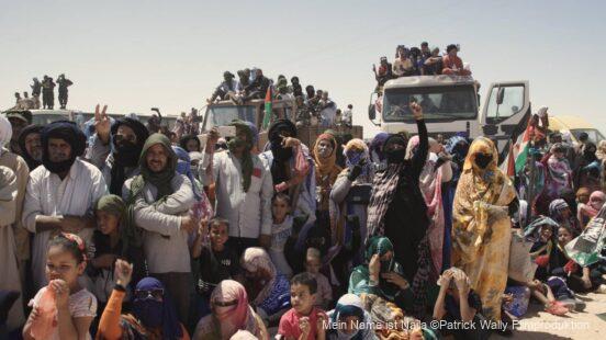 Eine große Gruppe von Menschen, darunter Männer, Frauen und Kinder in traditionellen, teils farbenfrohen Gewändern und Kopfbedeckungen, versammelt sich im Freien bei Tageslicht; im Hintergrund stehen und sitzen weitere Personen auf Lastern.