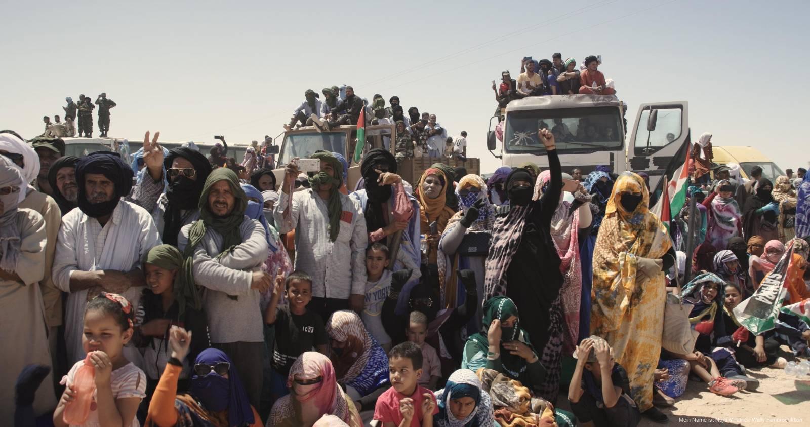 Eine große Gruppe von Menschen, darunter Männer, Frauen und Kinder in traditionellen, teils farbenfrohen Gewändern und Kopfbedeckungen, versammelt sich im Freien bei Tageslicht; im Hintergrund stehen und sitzen weitere Personen auf Lastern.