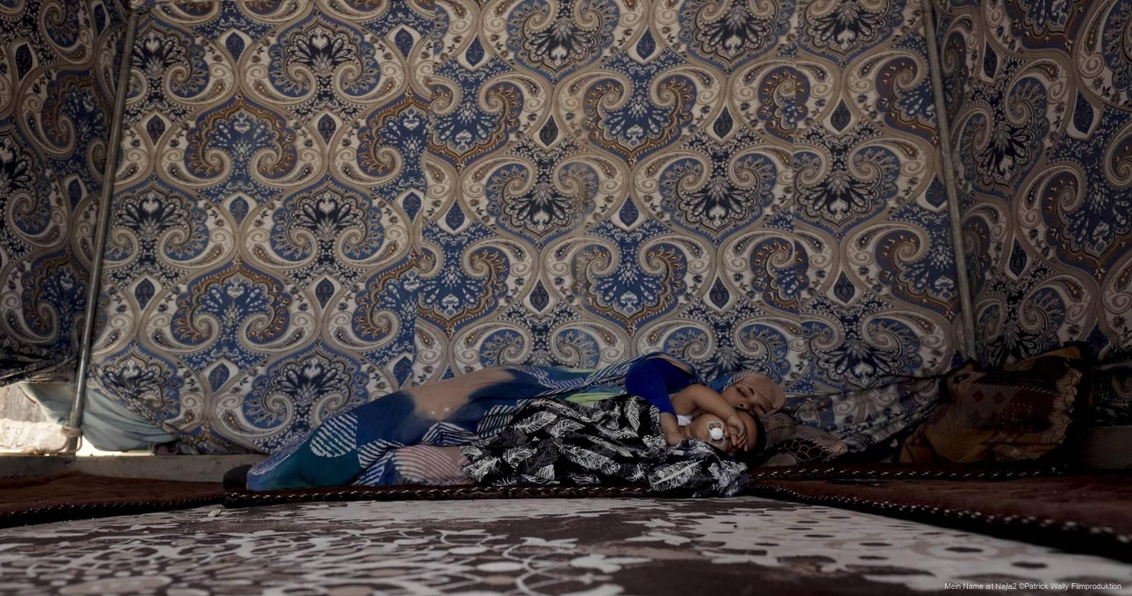 Ein Kind liegt auf einer Matratze in einem Zelt mit gemusterten, blauen Wandstoffen und schläft eng eingehüllt in Decken.