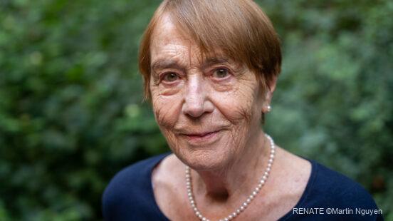 Ältere Frau mit kurzen braunen Haaren, Perlenkette und dunkelblauem Oberteil blickt leicht lächelnd in die Kamera, unscharfer grüner Hintergrund.