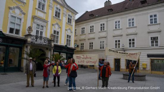 Gruppe von Menschen steht auf einem Platz vor historischen Gebäuden und hält Protestschilder; auf einem Schild steht "100 Jahre sind genug!" und auf dem anderen "Gehörlose können alles – außer hören!".