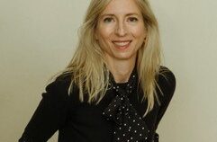 Frau mit langen blonden Haaren und schwarzer Bluse mit weißen Punkten lächelt vor neutralem Hintergrund.