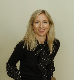 Frau mit langen blonden Haaren und schwarzer Bluse mit weißen Punkten lächelt vor neutralem Hintergrund.